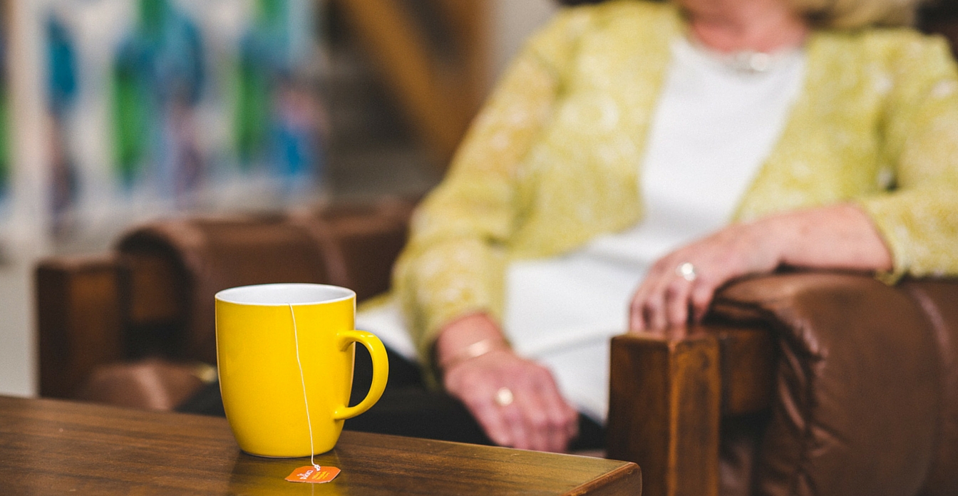 Close up of a yellow cup of tea with a woman sitting in an armchair in the background.