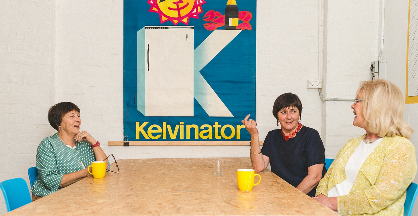 Three women in their 60s sitting around a table talking and laughing.